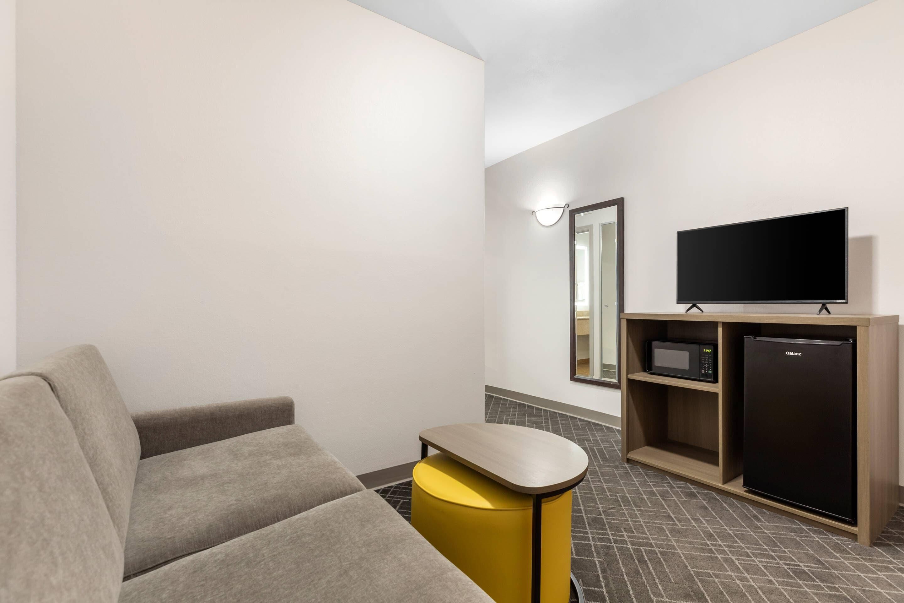 A modern hotel room with a sofa, a small yellow stool, a flat-screen TV, a microwave, and a refrigerator.