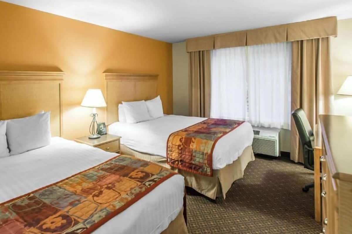 A hotel room with two beds, a desk, a chair, a lamp, and a window with curtains.