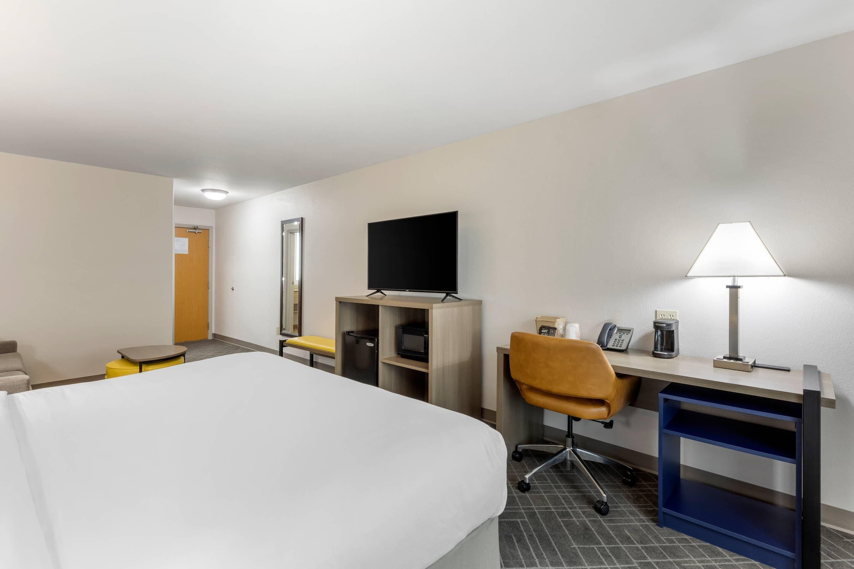 A hotel room with a bed, desk, chair, TV, lamp, and a small table with a coffee maker.
