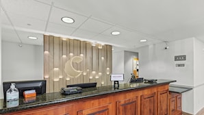 Un área de recepción con un mostrador de madera, un letrero de bienvenida, una pantalla digital y una cámara en el techo.