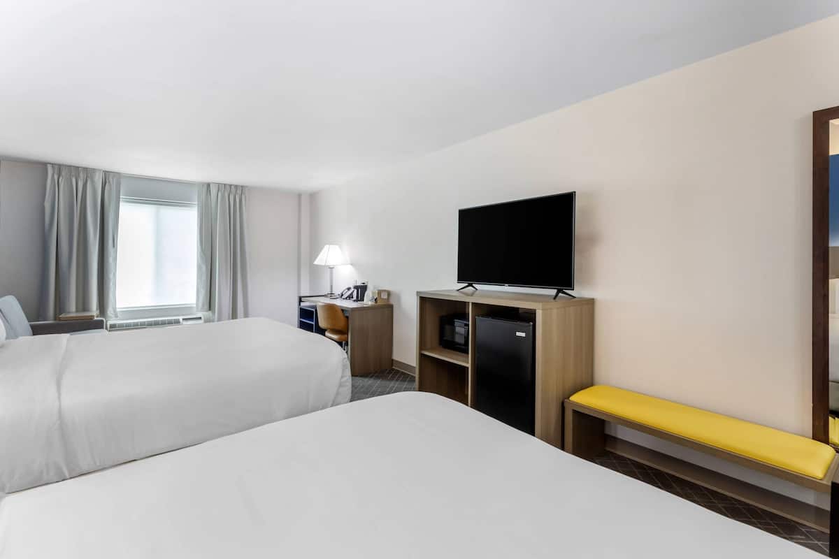 A hotel room with two beds, a desk, a television, and a bench.