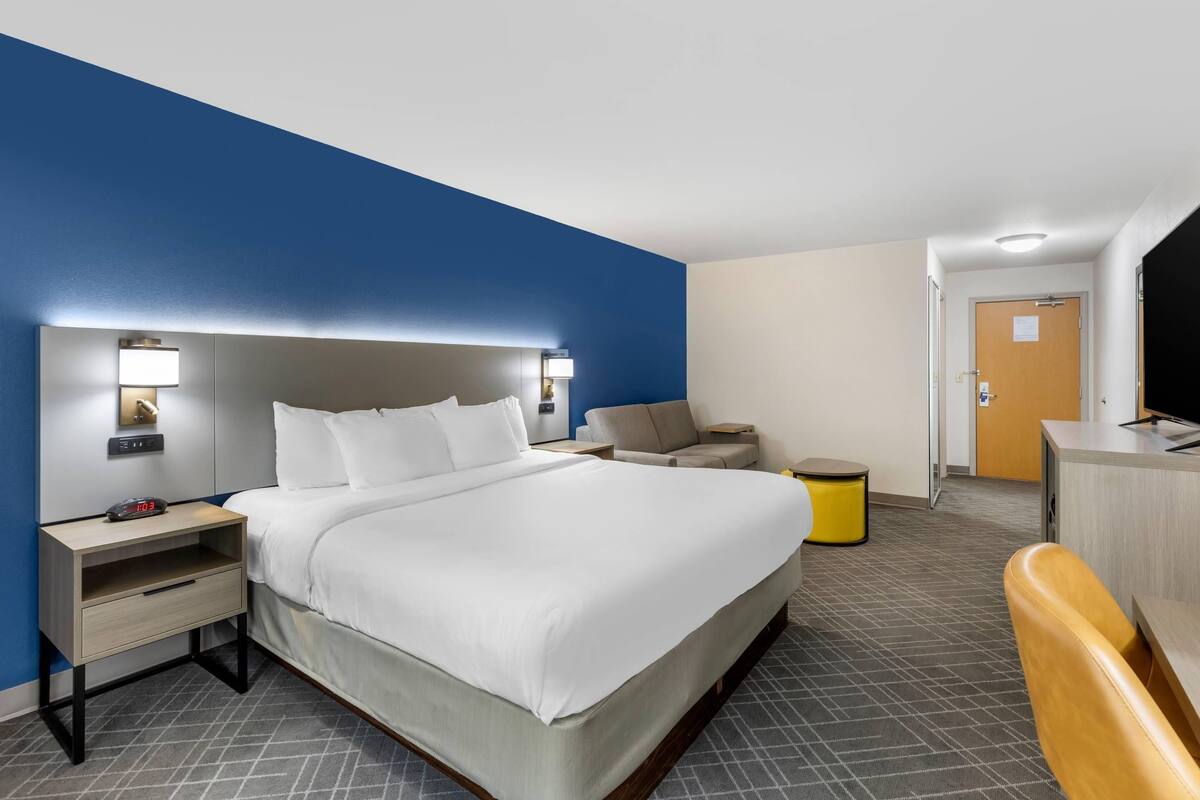 A hotel room with a bed, bedside tables, a desk, a chair, and a small stool.