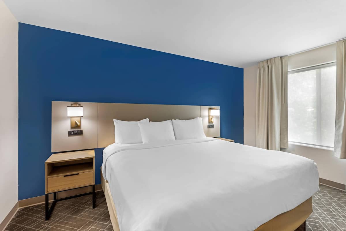 A hotel room with a large bed, a nightstand, a wall-mounted lamp, and a window with curtains.