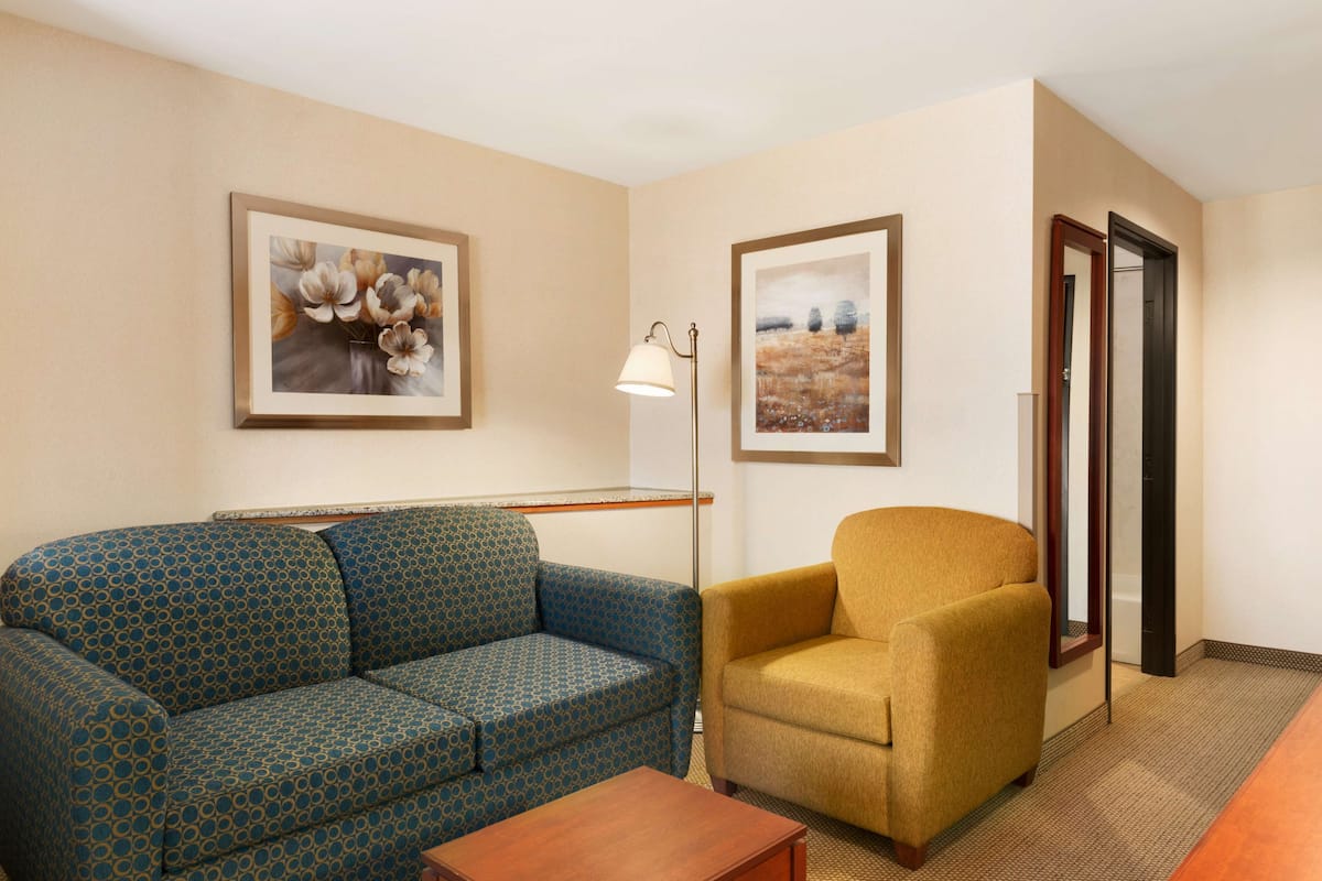 A room with a patterned sofa, a single armchair, a floor lamp, and two framed pictures on the wall.