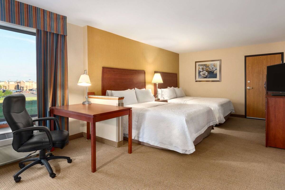 A hotel room with two beds, a desk, a chair, a TV, and a painting on the wall.