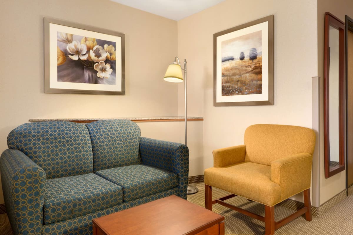 A living room with a patterned sofa, a yellow armchair, a floor lamp, and two framed artworks on the walls.