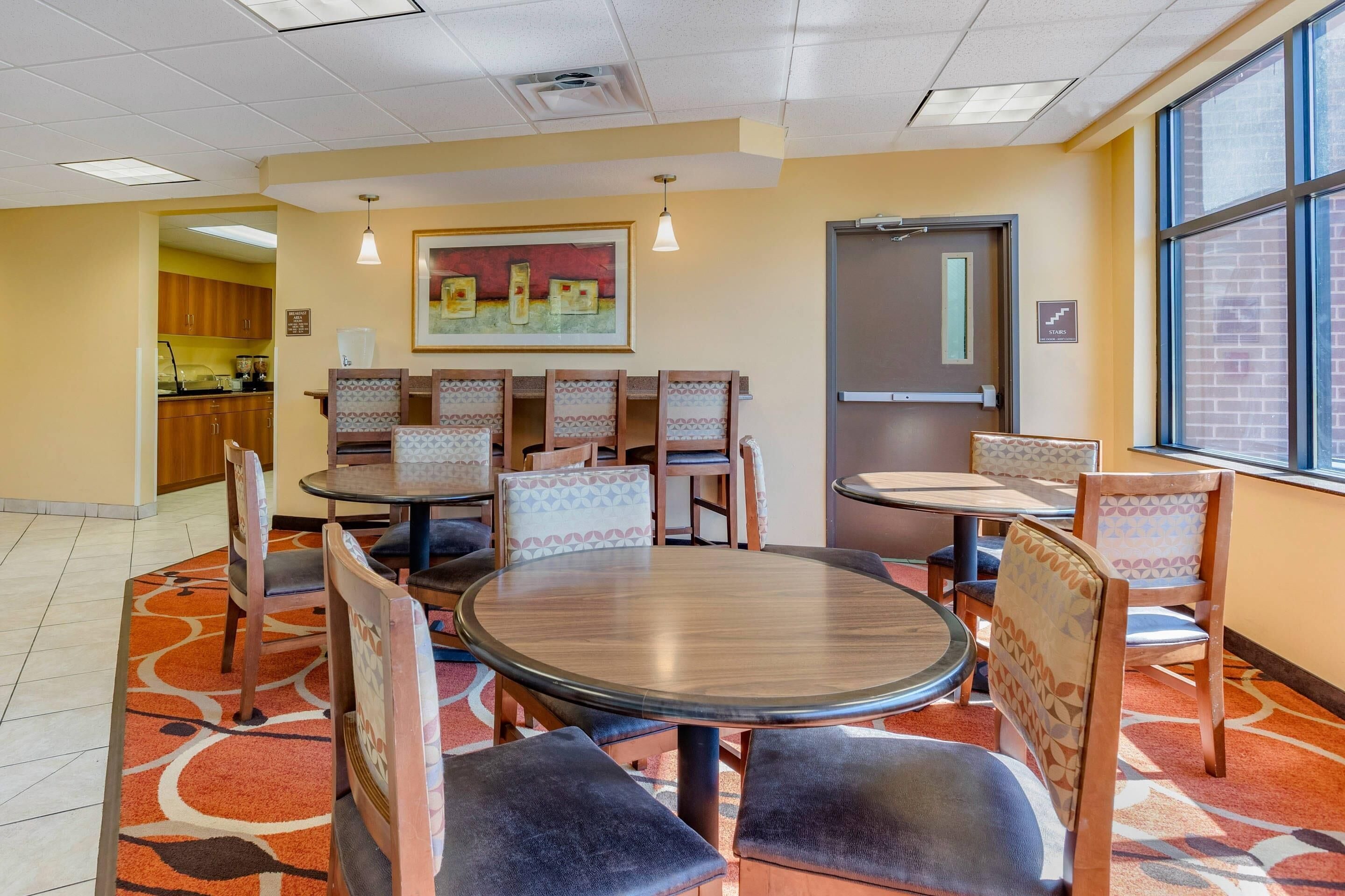 A dining area with round tables and chairs, a painting on the wall, and a view through a window.