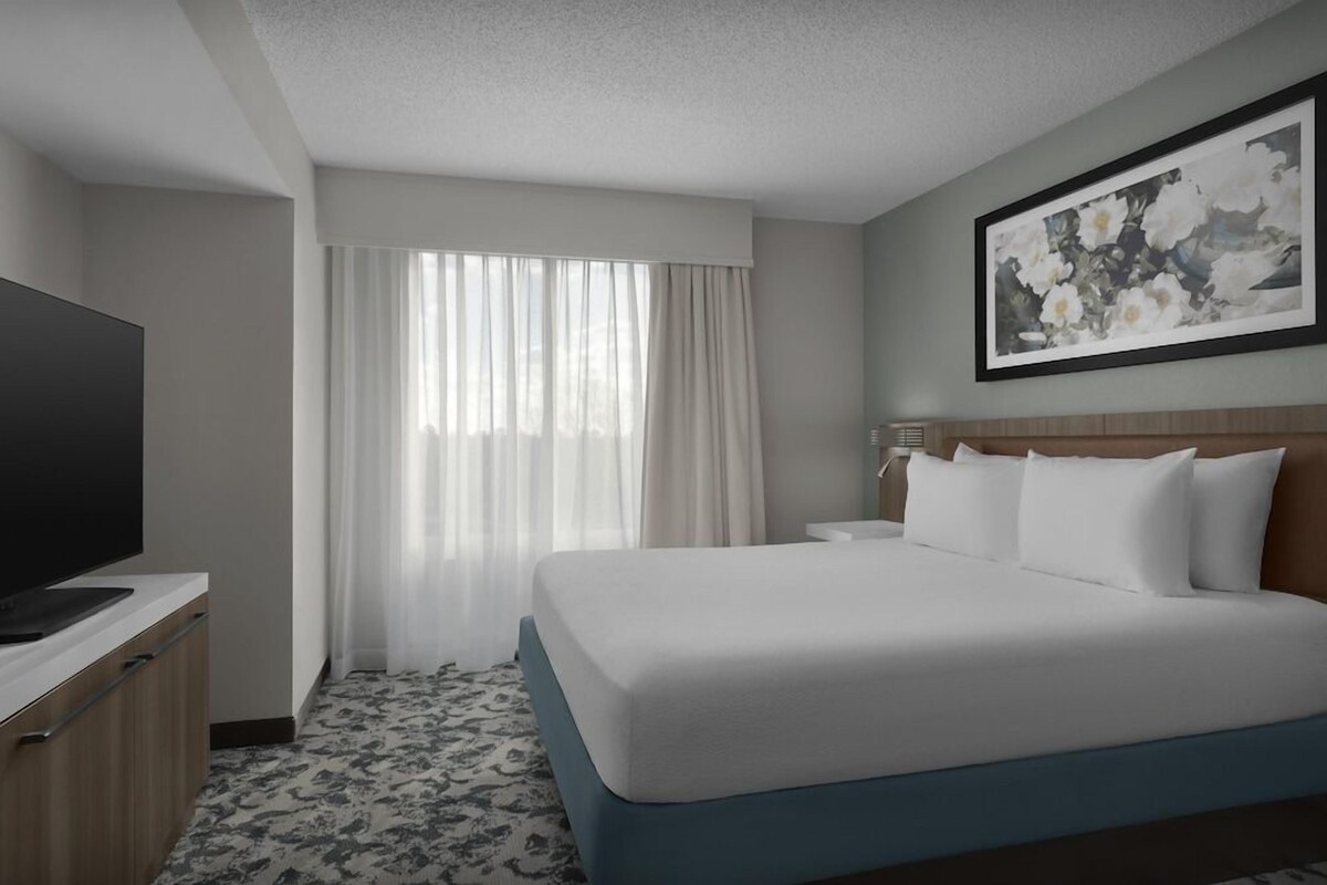 A hotel room with a bed, a TV, a desk, and a painting on the wall.