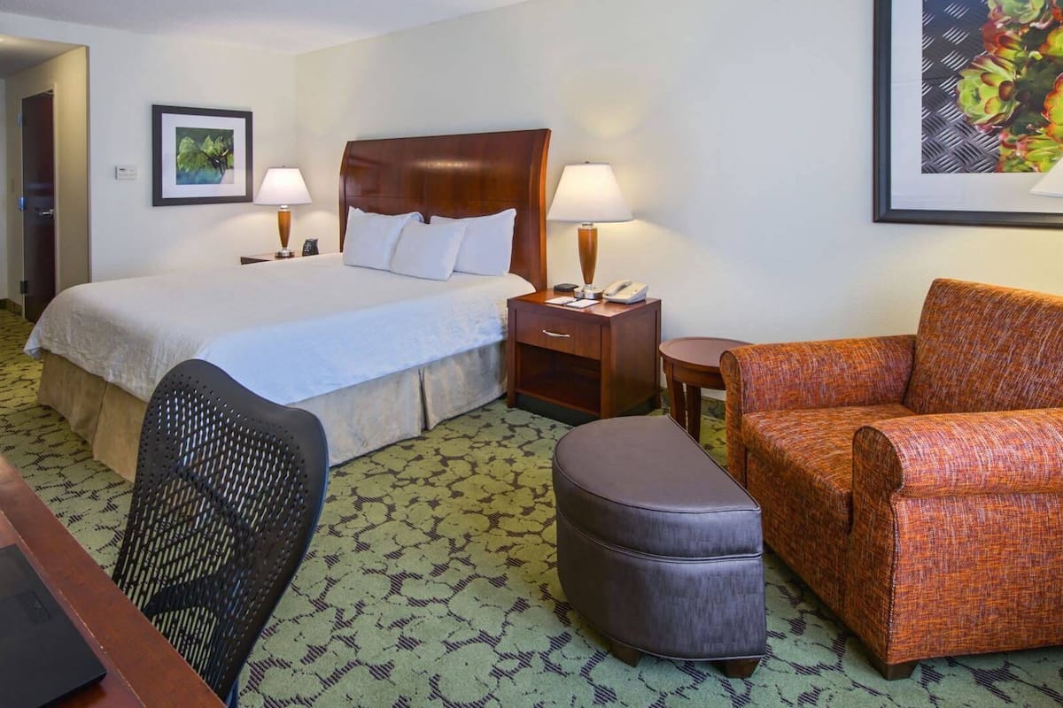 A hotel room with a bed, two bedside tables, a desk with a laptop, a chair, an ottoman, and a sofa.