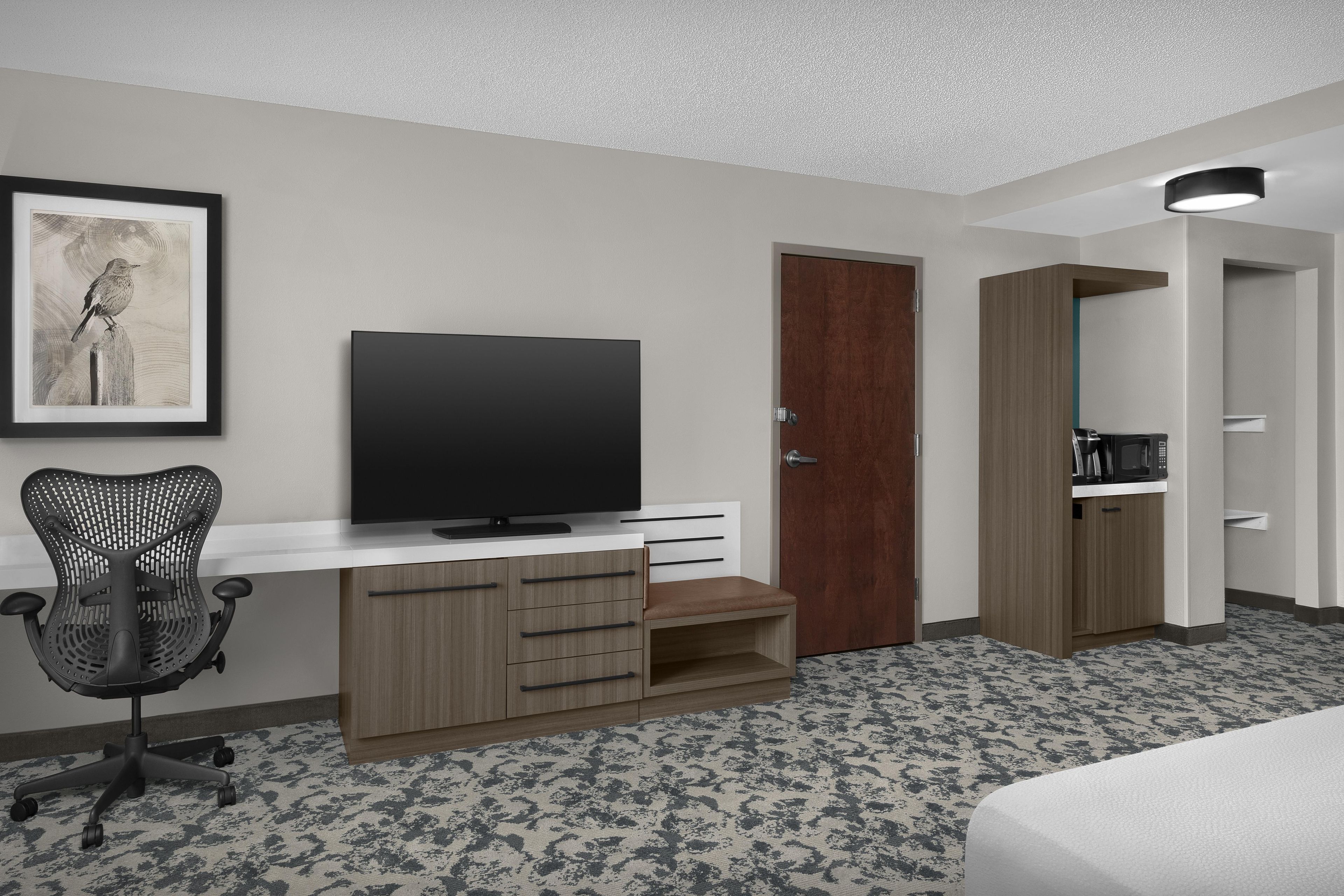 Habitación de hotel con televisor de pantalla plana, escritorio de madera, silla de oficina negra, armario de madera y un cuadro en la pared.
