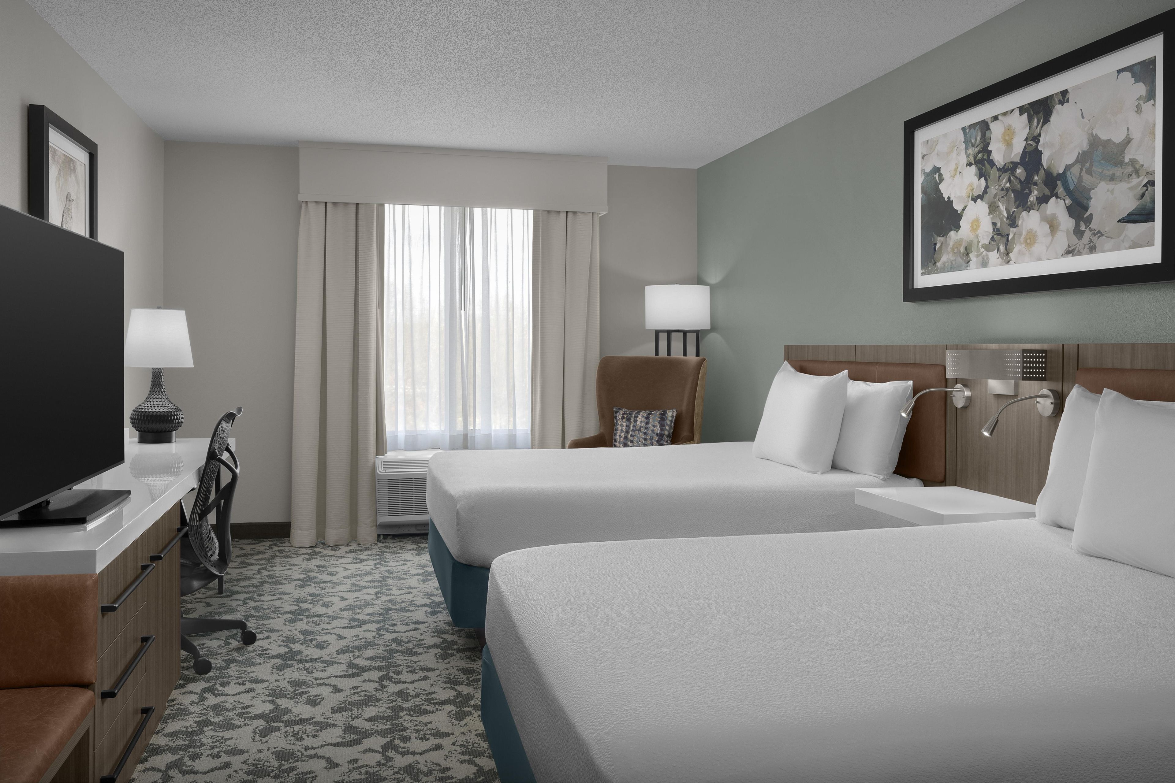 A hotel room with two beds, a desk with a TV, a lamp, and a painting on the wall.