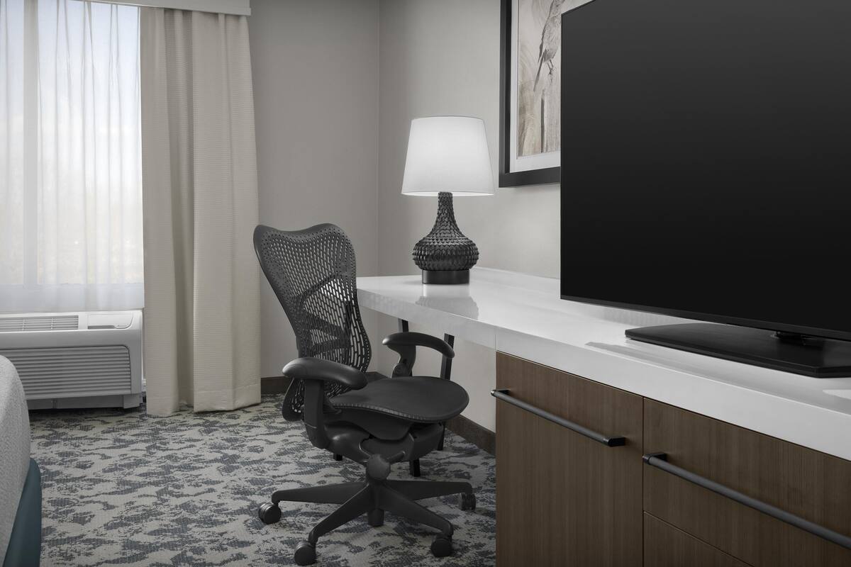 A hotel room with a bed, a desk with a TV, a chair, a lamp, and a window with curtains.