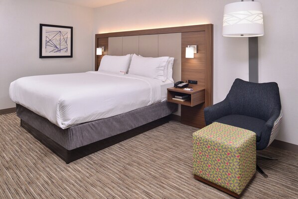 A hotel room with a bed, bedside table, lamp, chair, and a small ottoman.