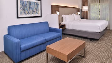 A hotel room with a blue sofa, a wooden coffee table, a bed with white bedding, and a painting on the wall.