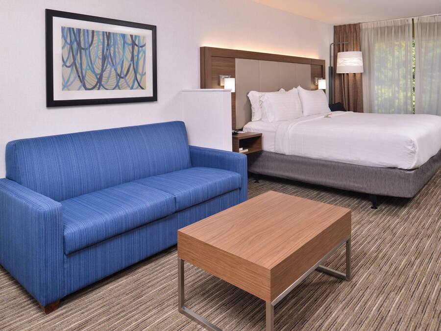 A hotel room with a blue sofa, a wooden coffee table, a bed with white bedding, and a painting on the wall.