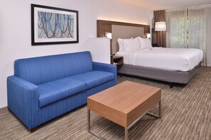 A hotel room with a blue sofa, a wooden coffee table, a bed with white bedding, and a painting on the wall. - Holiday Inn Express & Suites Lacey – Olympia by IHG (Lacey)
