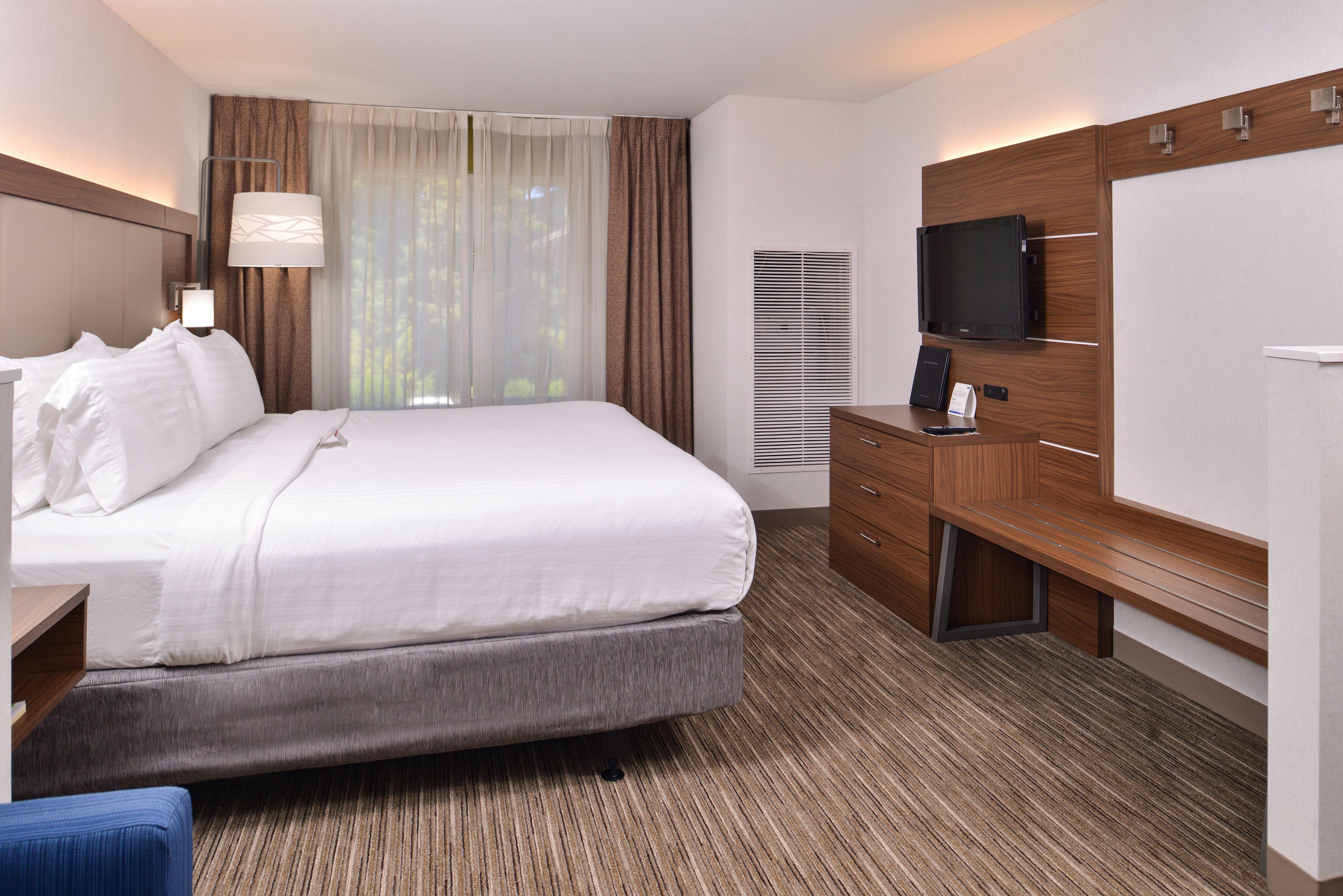 A hotel room with a large bed, a desk with a flat-screen TV, a wooden dresser, and a window with curtains.
