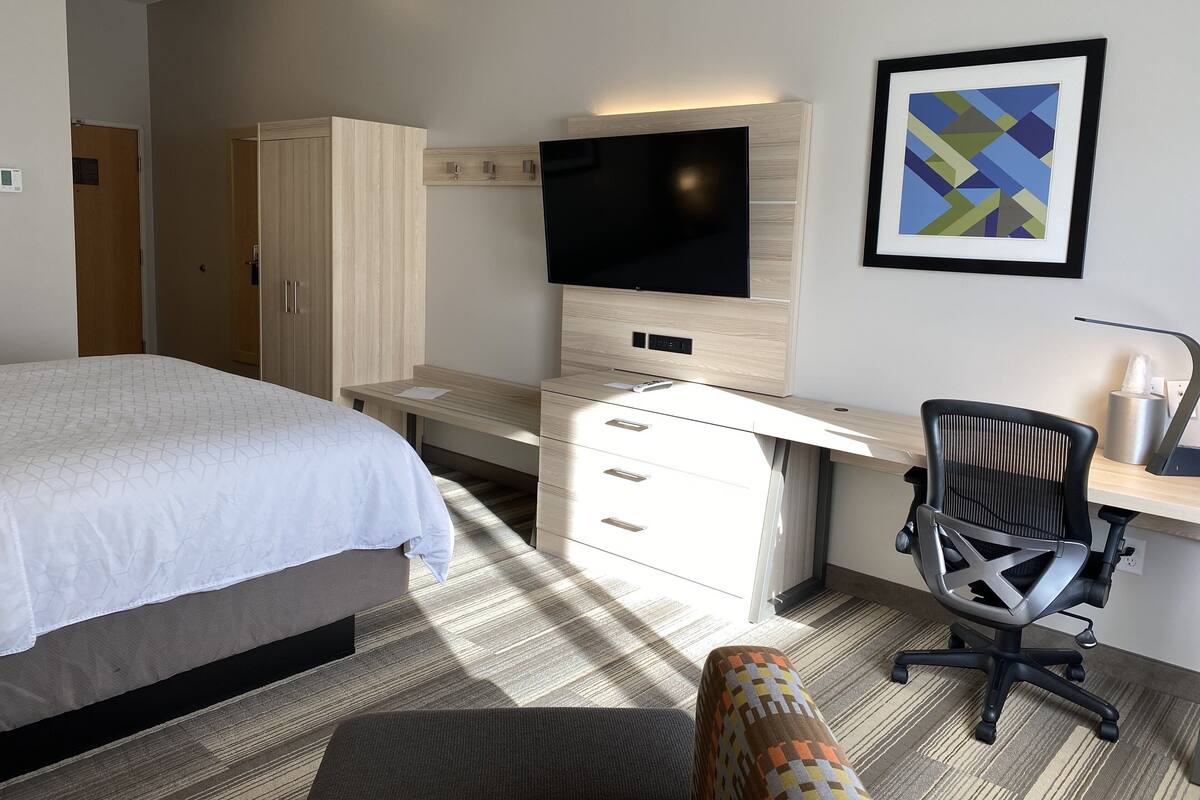 A hotel room with a bed, a desk with a computer, a chair, a TV, and a framed picture on the wall.