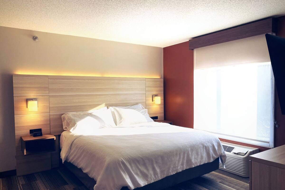 A hotel room with a bed, bedside tables, a nightstand, a heater, and a window with curtains.