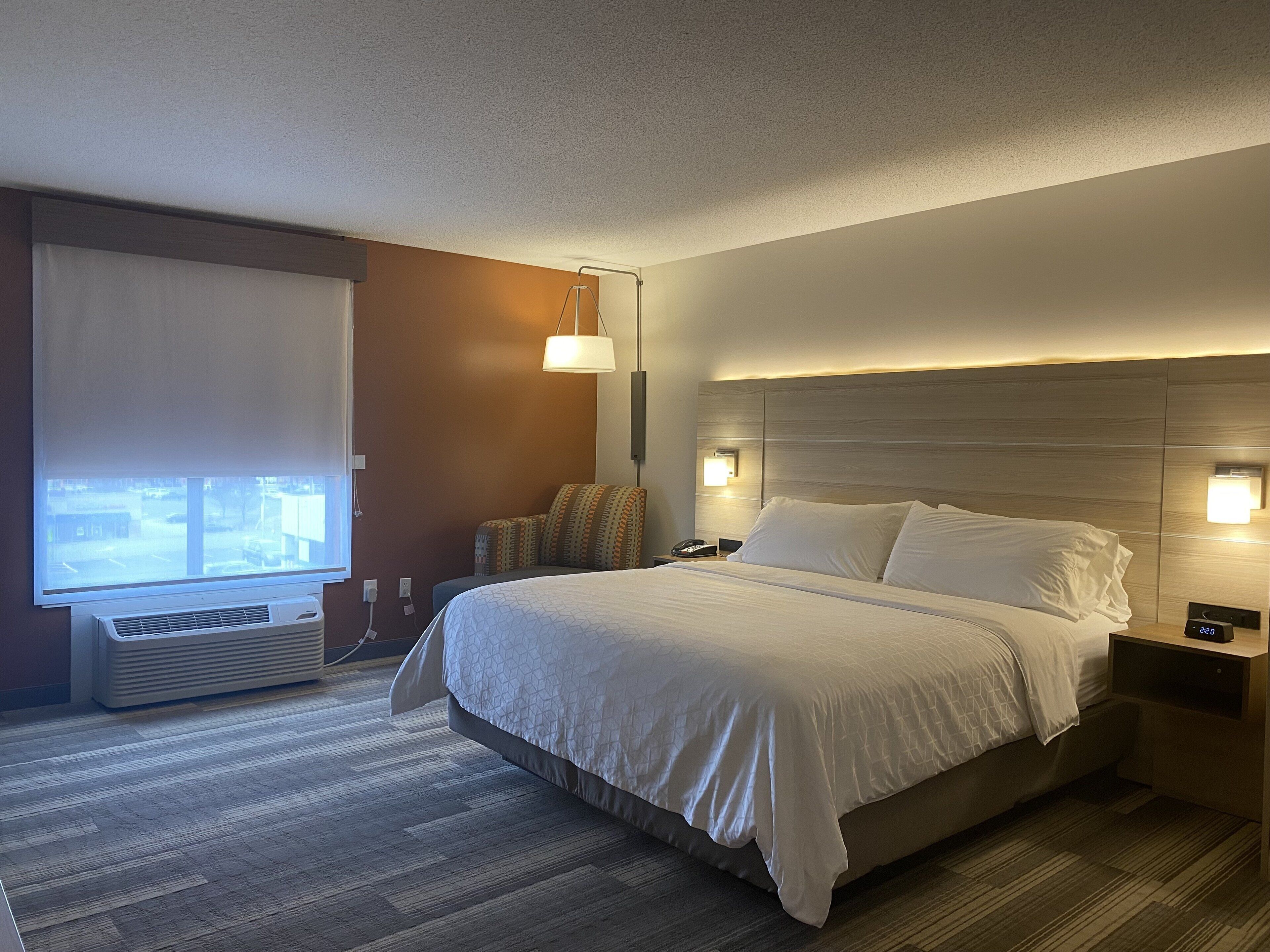 A hotel room with a large bed, bedside lamps, a window with blinds, and a wall-mounted air conditioner.