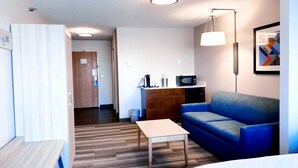 Une chambre d’hôtel moderne avec un plancher en bois, un canapé bleu, une table en bois, un lit, une kitchenette équipée d’un four à micro-ondes et un luminaire au plafond.