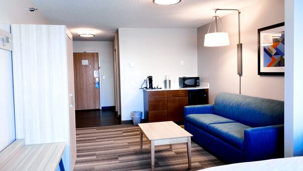 A modern hotel room with a wooden floor, a blue sofa, a wooden table, a bed, a kitchenette with a microwave, and a ceiling light.