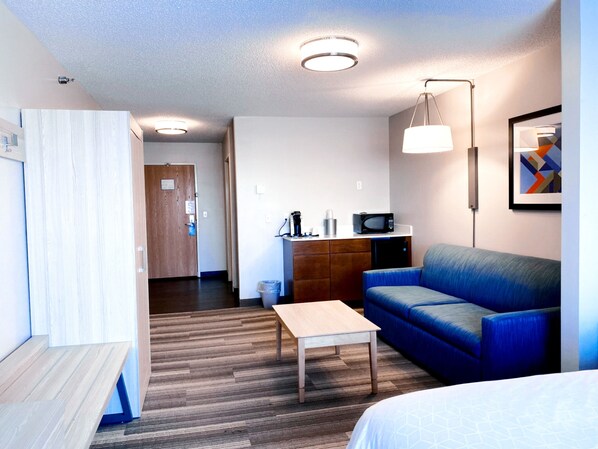 A modern hotel room with a wooden floor, a blue sofa, a wooden table, a bed, a kitchenette with a microwave, and a ceiling light. - Holiday Inn Express Hotel & Suites Logansport by IHG (Logansport)