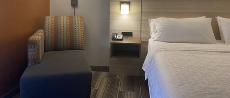 A hotel room with a bed, a bedside table with a phone, a lamp, and a patterned chair.