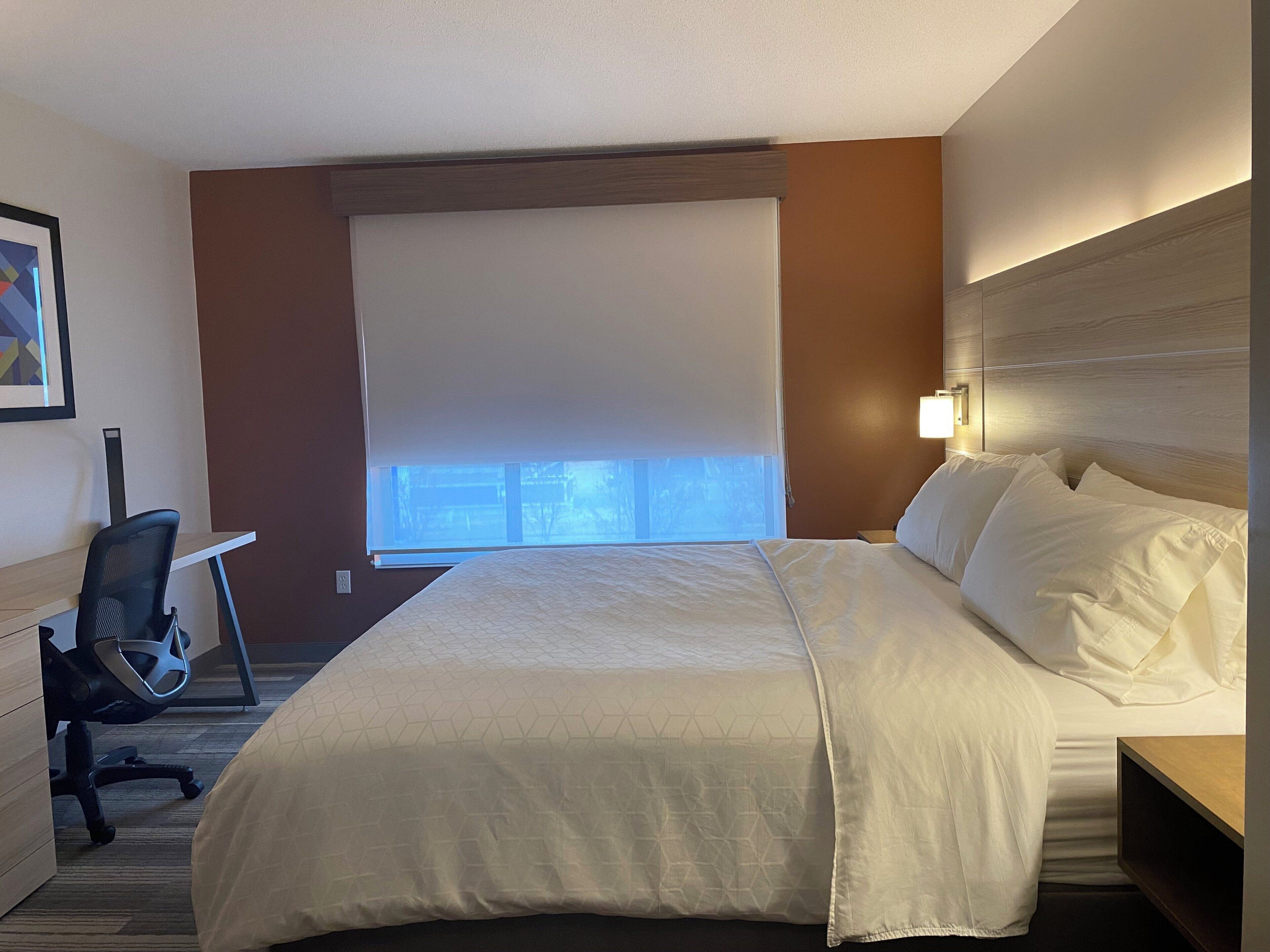 A hotel room with a bed, desk, chair, and a window with blinds.