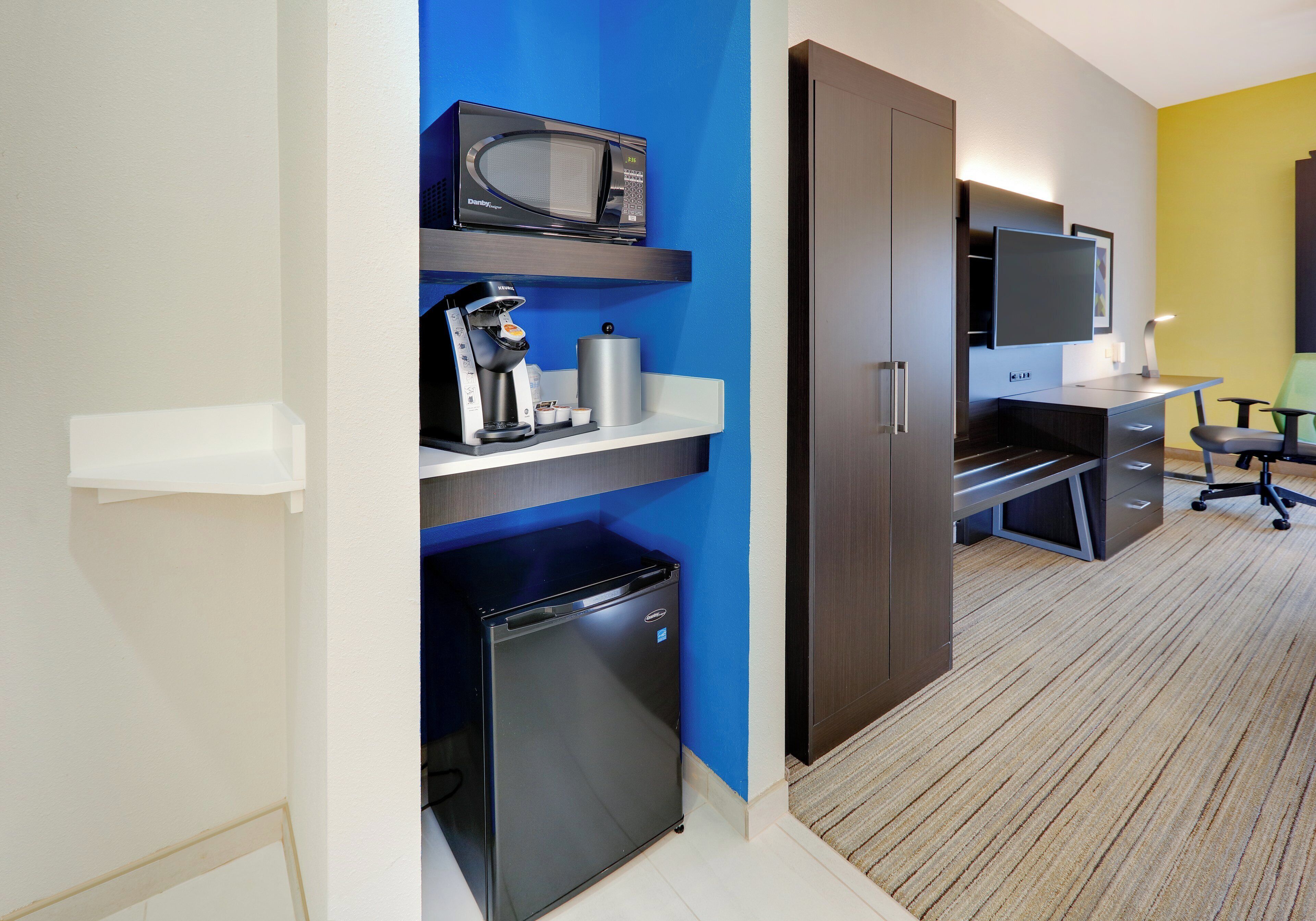 A hotel room with a kitchenette, a microwave, a refrigerator, a television, and a desk with a chair.