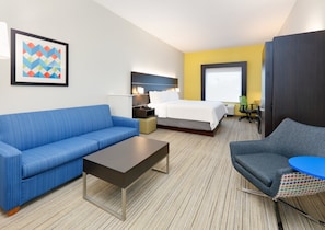 A hotel room with a blue sofa, a grey armchair, a small table, a bed with white linens, a nightstand with a lamp, and a colorful abstract painting on the wall. - Holiday Inn Express Hotel & Suites Cleburne by IHG (Cleburne)