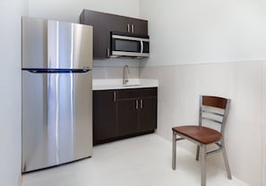 A compact kitchen with a stainless steel refrigerator, a microwave, dark wooden cabinets, a white countertop, and a metal chair. - Holiday Inn Express Hotel & Suites Cleburne by IHG (Cleburne)
