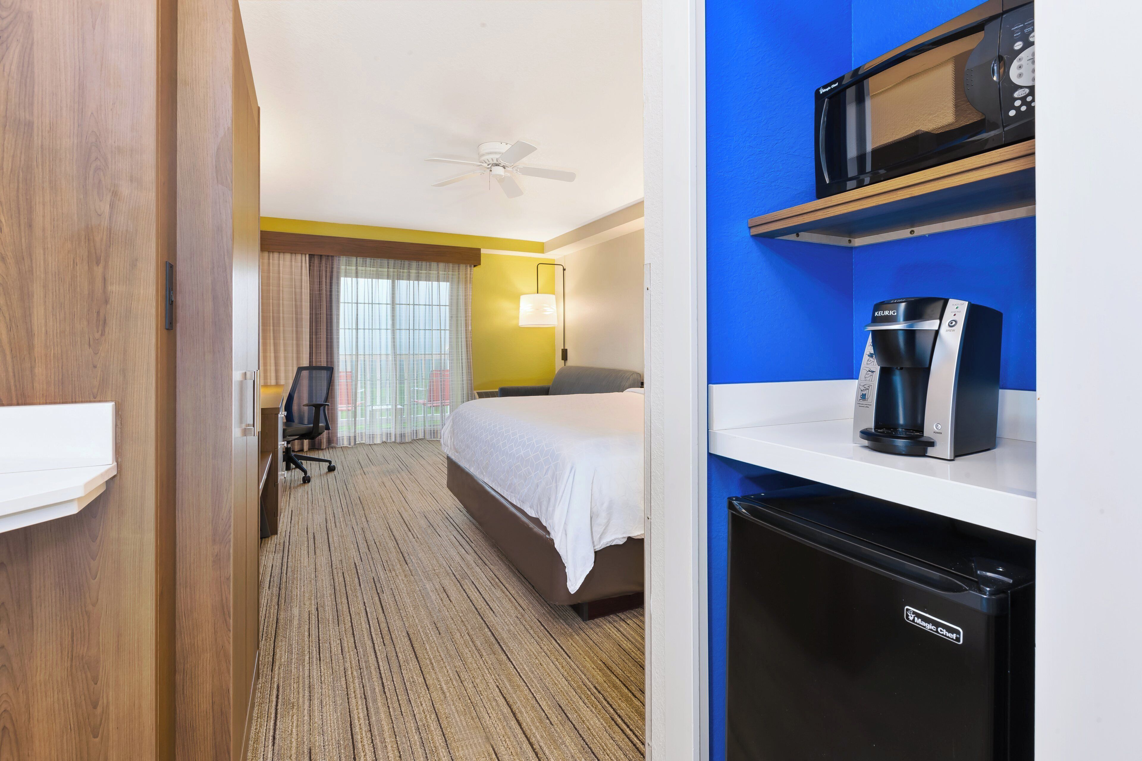Habitación de hotel con cama, escritorio, silla, microondas, cafetera y refrigerador.