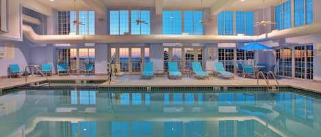 A large indoor swimming pool with multiple lanes, lounge chairs, and a view of the sunset through the windows.