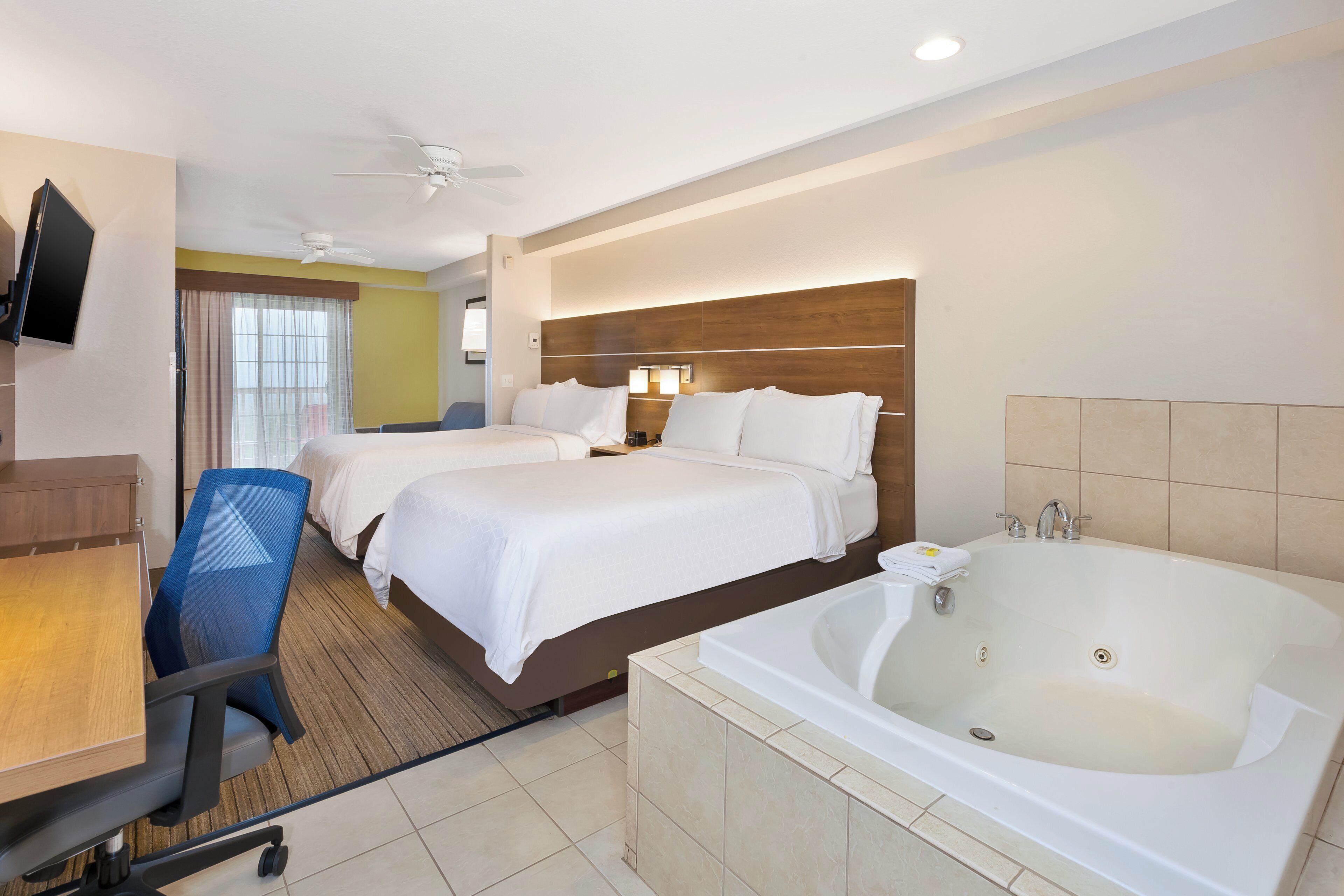 A hotel room with two beds, a desk with a chair, a television, a ceiling fan, and a large bathtub.