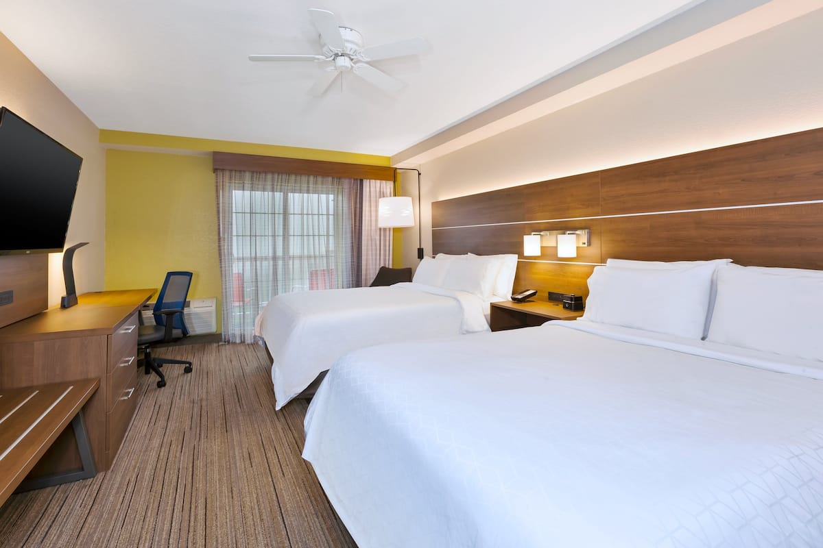 Habitación de hotel con dos camas, un escritorio, una silla, un televisor, un ventilador de techo y una ventana con cortinas.