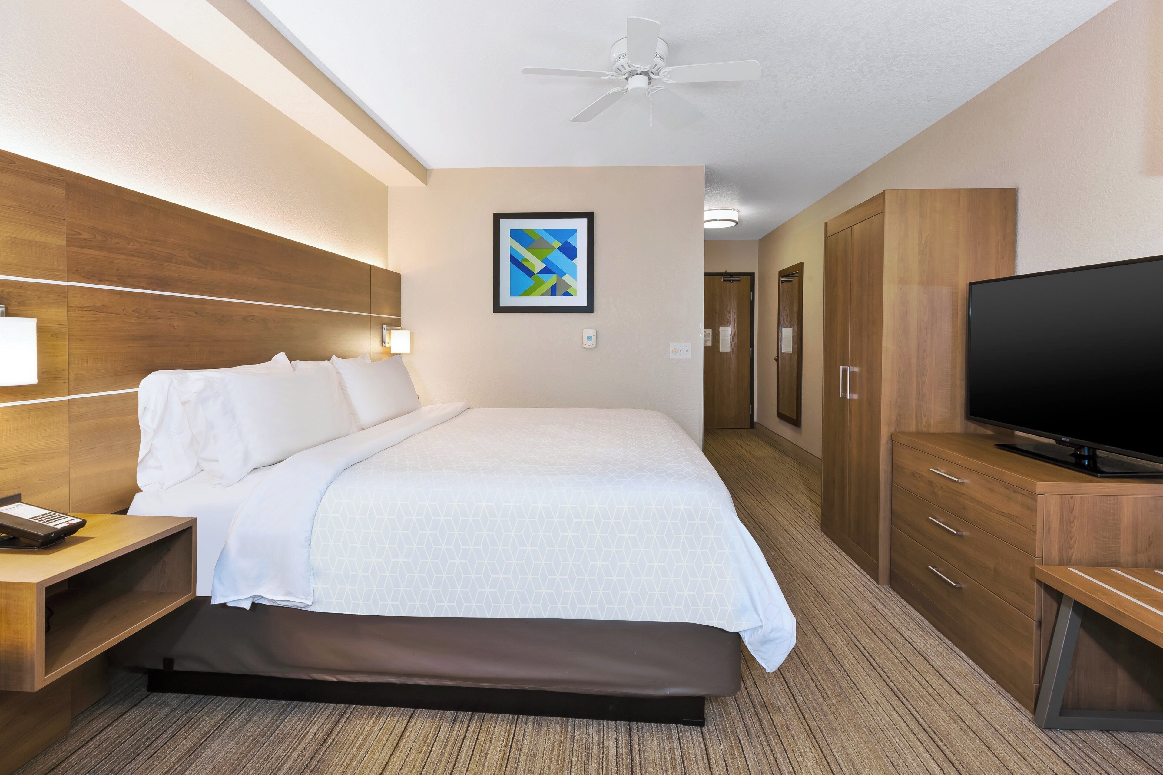 A hotel room with a bed, a television, a desk with a phone, a wardrobe, and a ceiling fan.