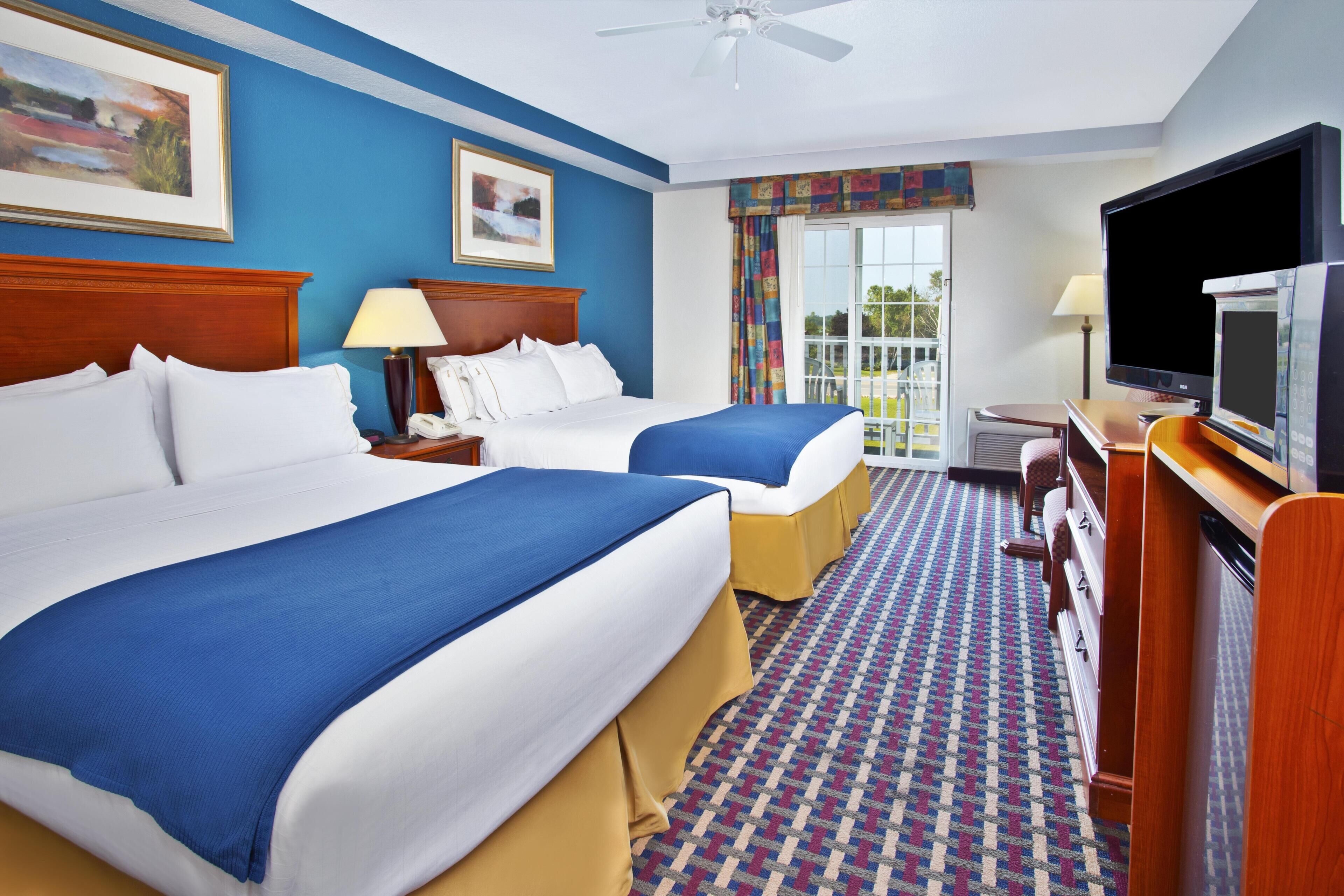 A hotel room with two beds, a television, a desk, and a view of the outdoors.