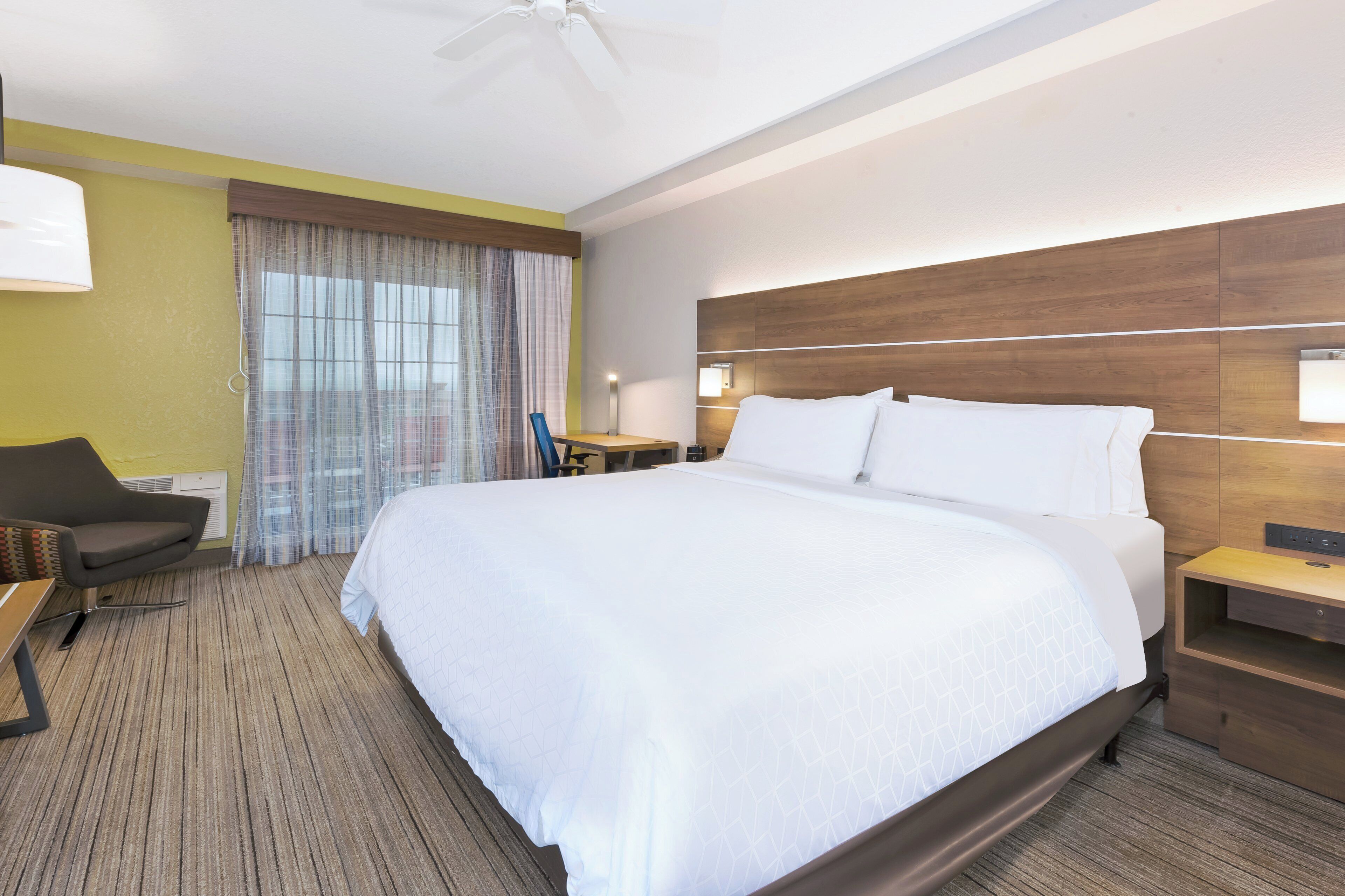 A hotel room with a large bed, a desk, a chair, a nightstand, and a window with curtains.