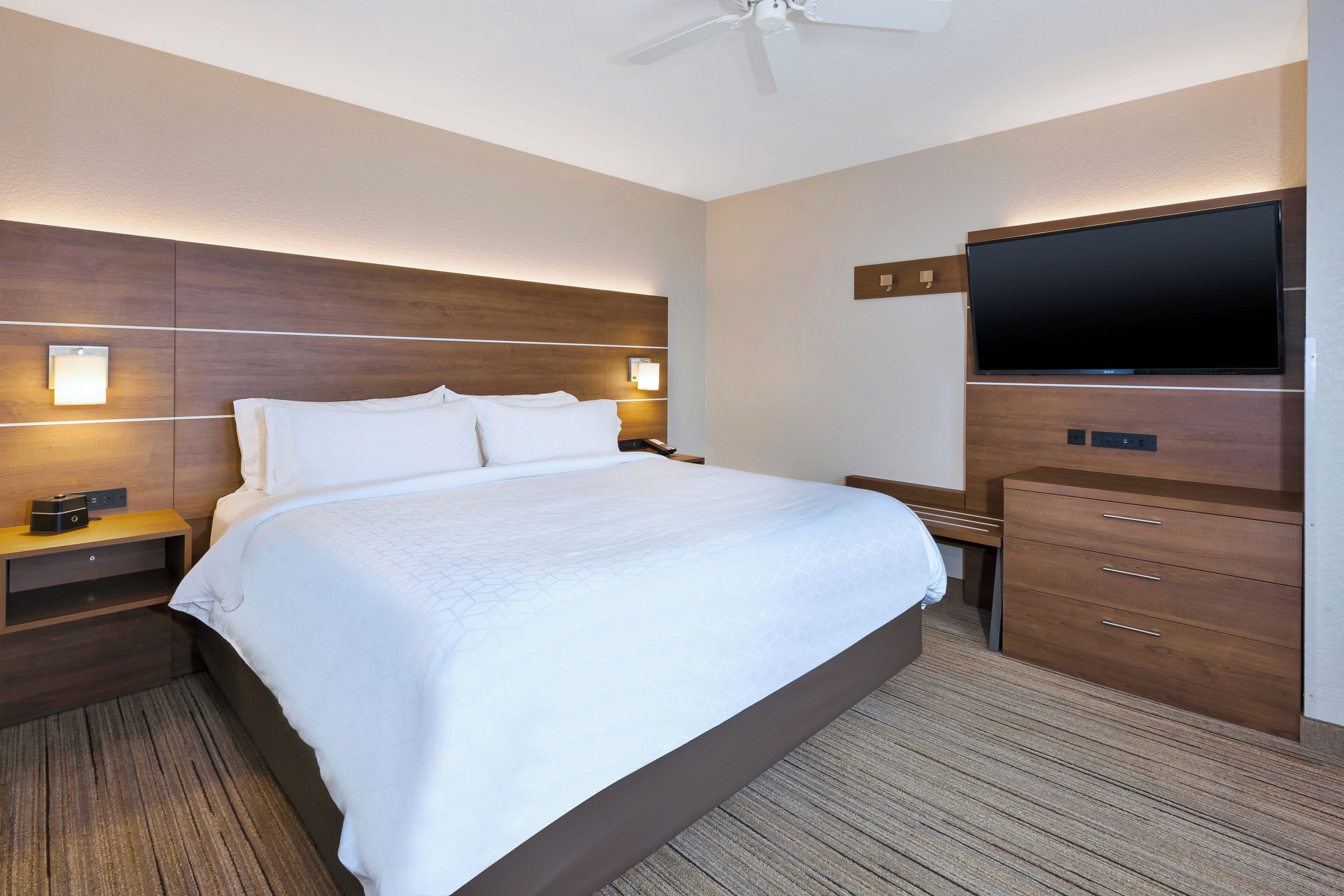 A hotel room with a large bed, a flat-screen TV, wooden headboard, bedside tables, and a carpeted floor.