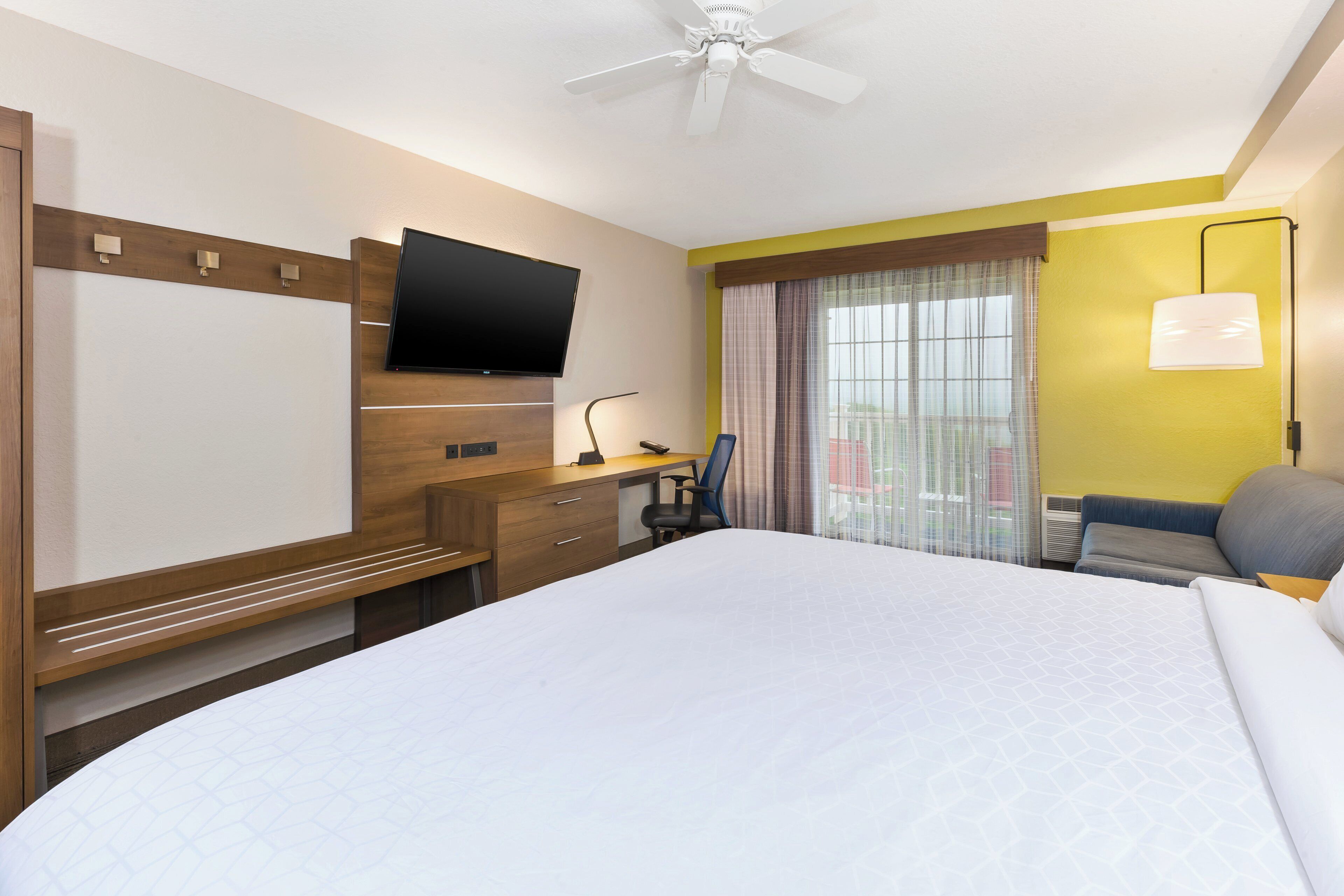 Habitación de hotel con cama, escritorio, silla, televisor y ventana con cortinas.