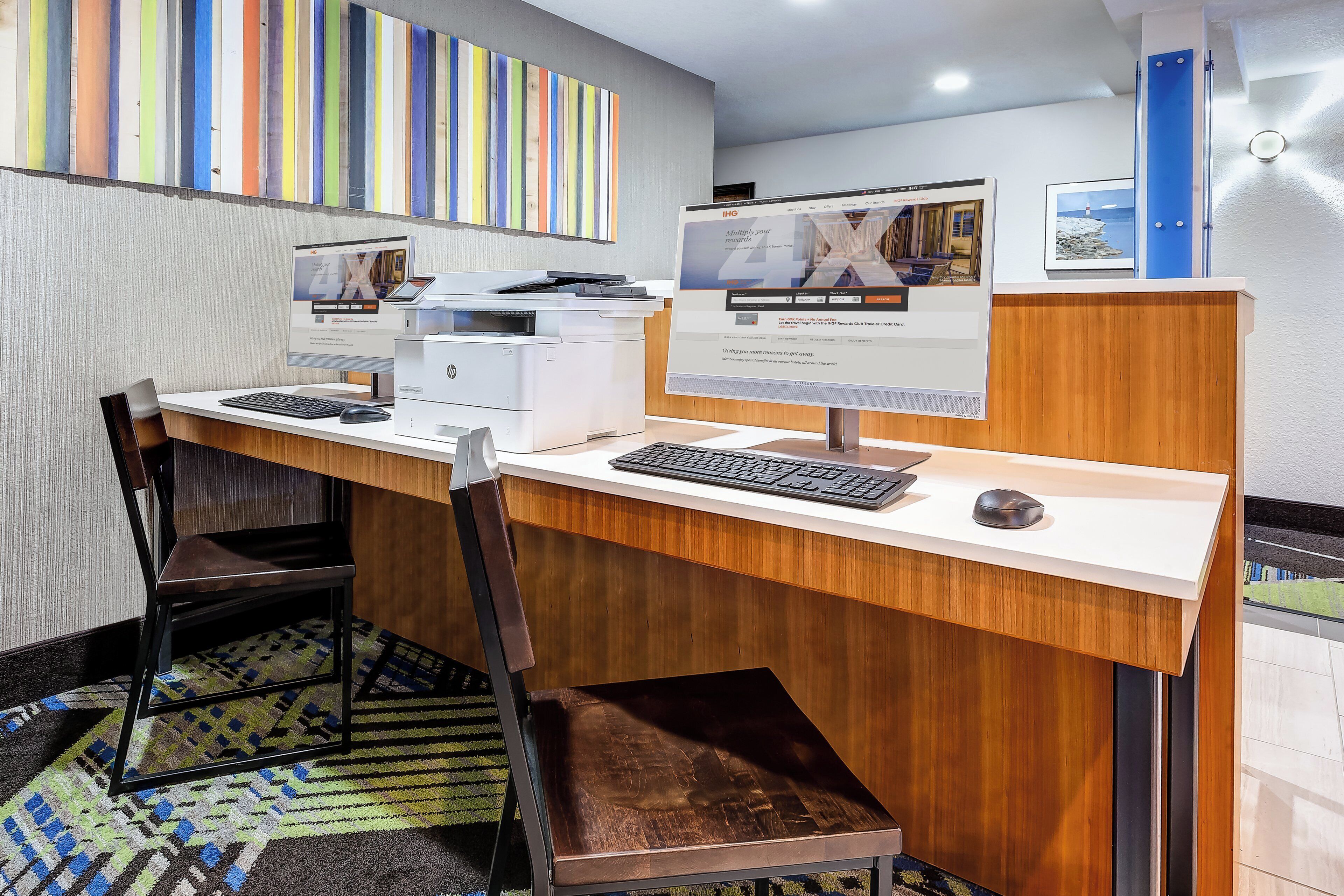 A hotel room with a desk, two chairs, a printer, and a computer monitor.