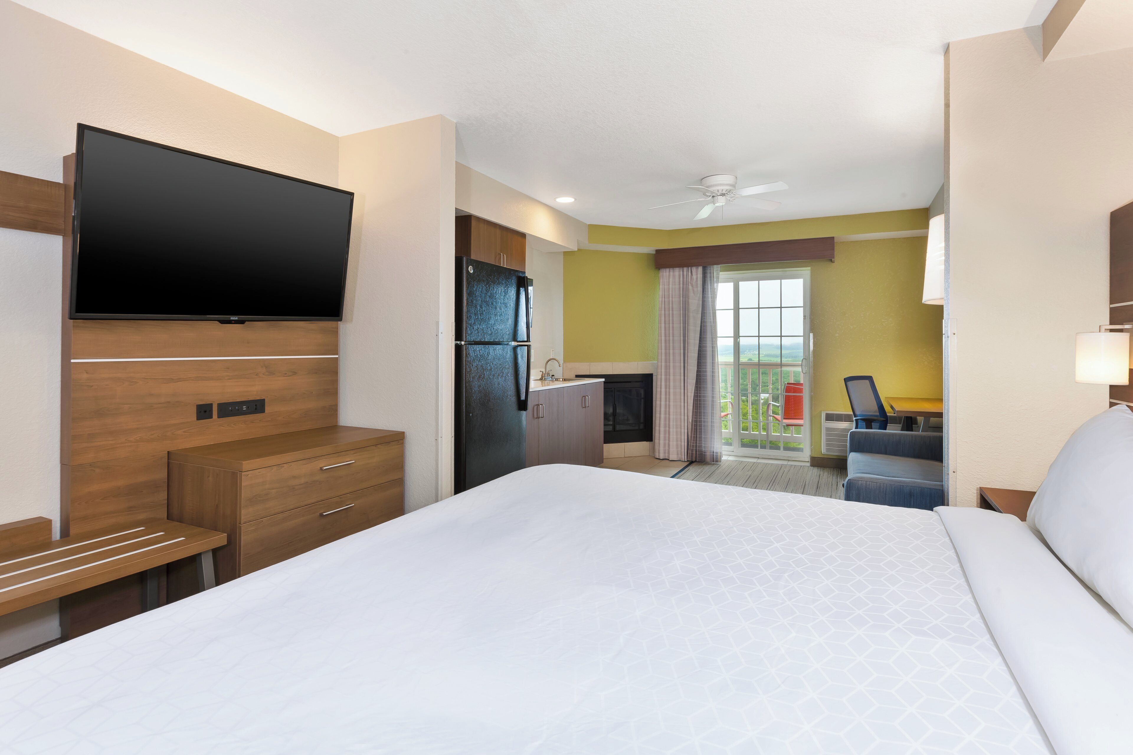A hotel room with a bed, a flat-screen TV, a kitchenette, and a balcony with a view.