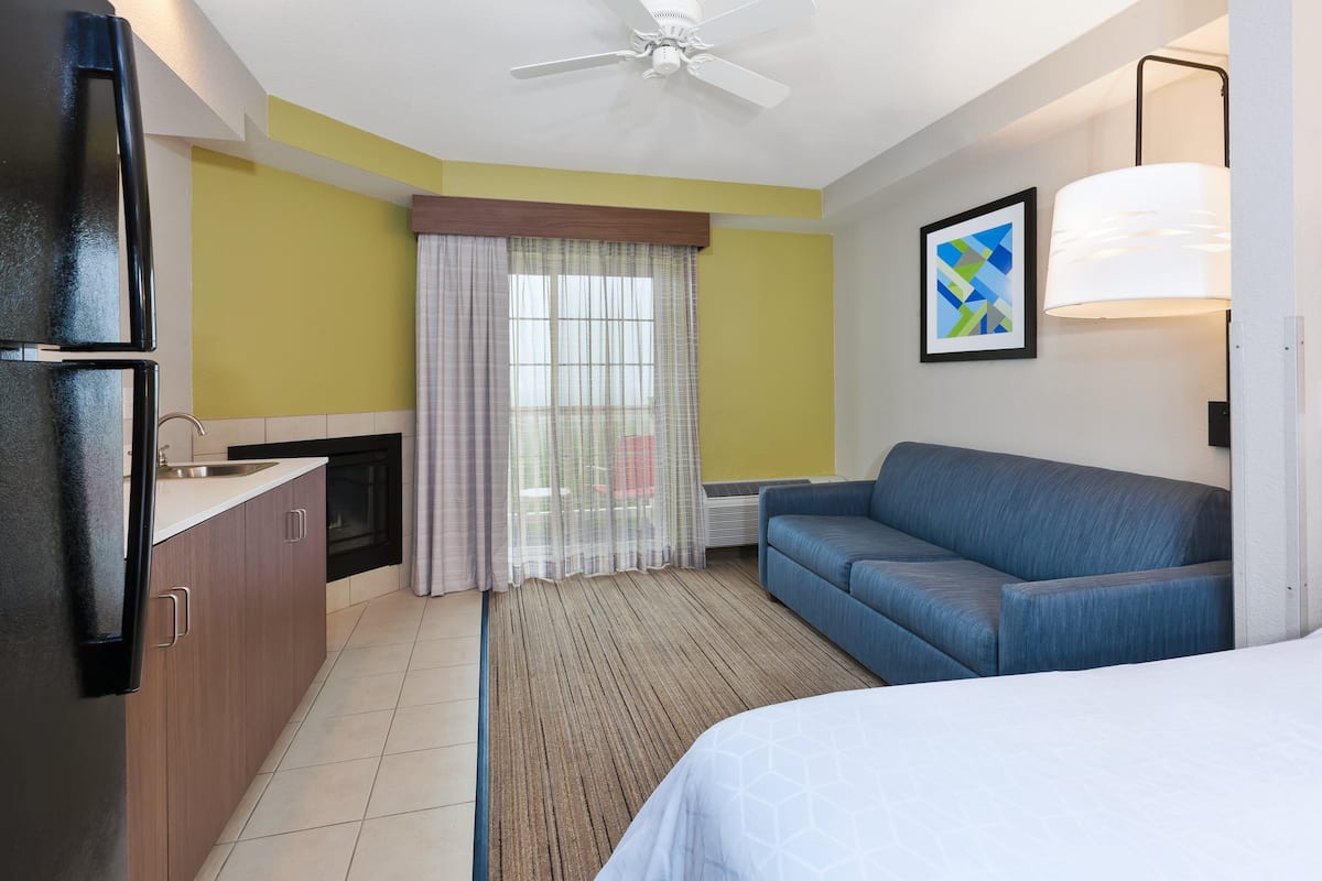Habitación de hotel con kitchenette, sofá, cama, ventilador de techo y ventana con cortinas.