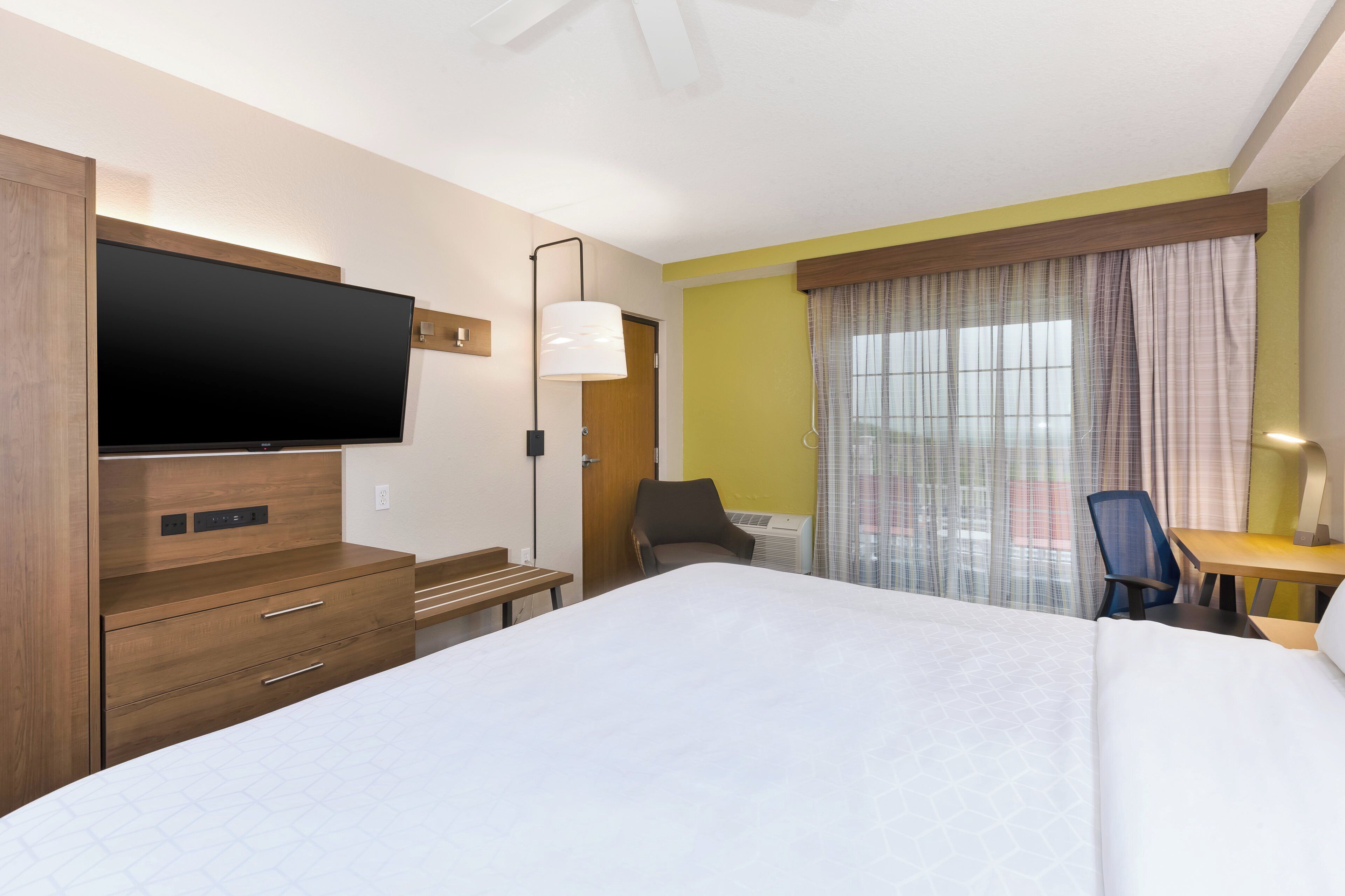 Una habitación de hotel con una cama grande, un televisor de pantalla plana, un escritorio con silla y una ventana con cortinas.