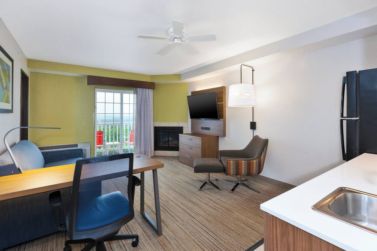 Habitación de hotel con escritorio, silla, televisor, kitchenette y vistas al exterior.