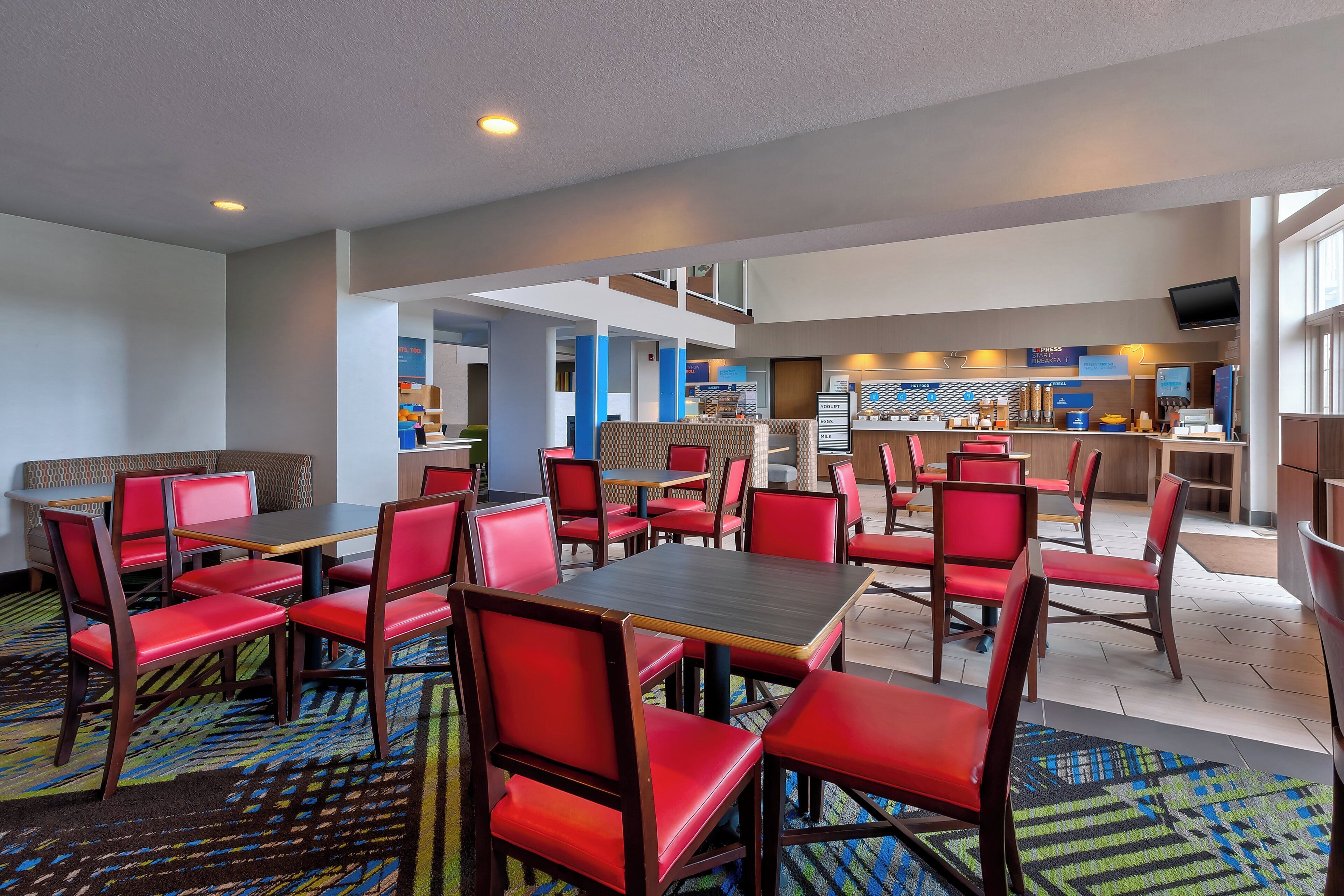 A hotel restaurant with multiple tables and chairs, a counter with food and drink options, and a seating area with a sofa and chairs.