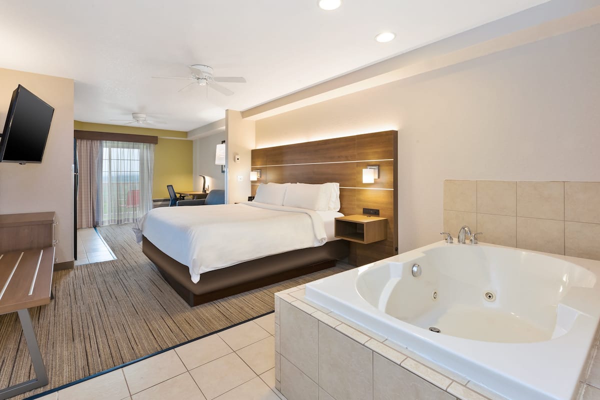 Una habitación de hotel con una cama grande, un espacio de descanso con una mesa y sillas, un baño independiente con bañera y un televisor montado en la pared.