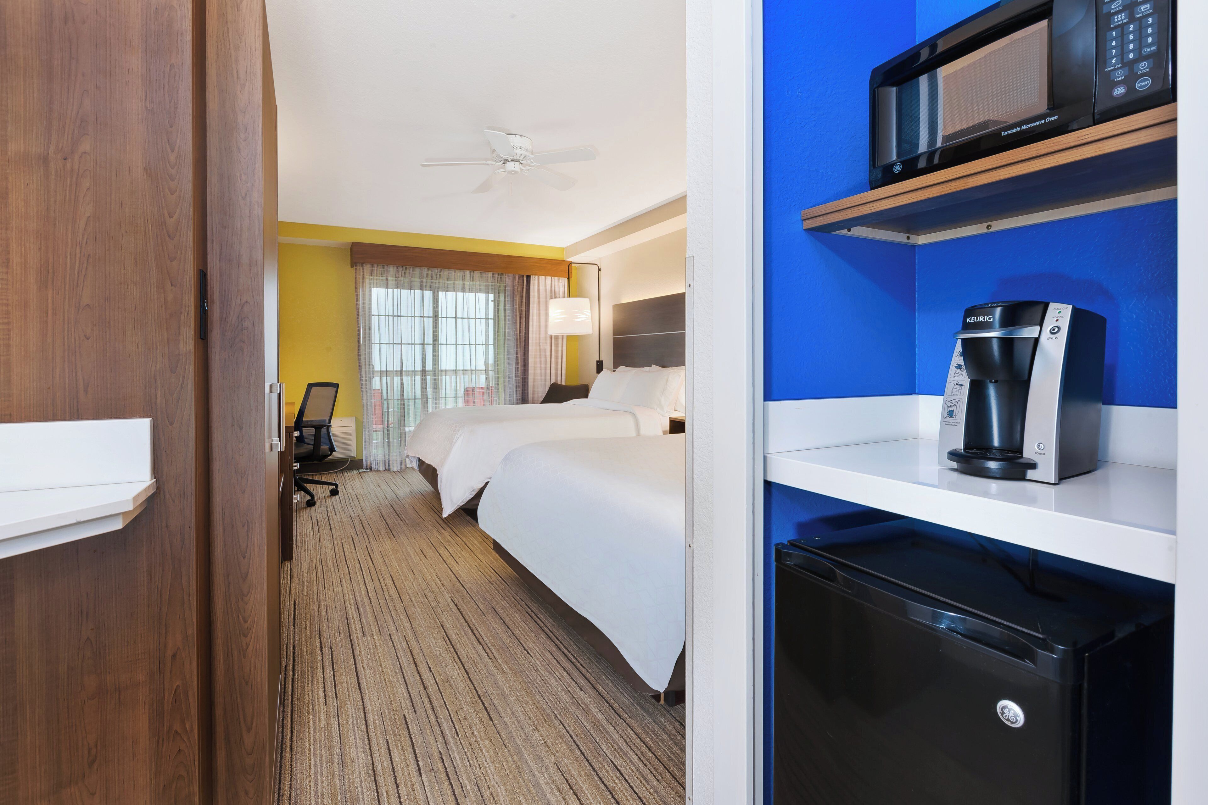 A hotel room with a bed, a desk, a chair, a microwave, a coffee maker, and a refrigerator.