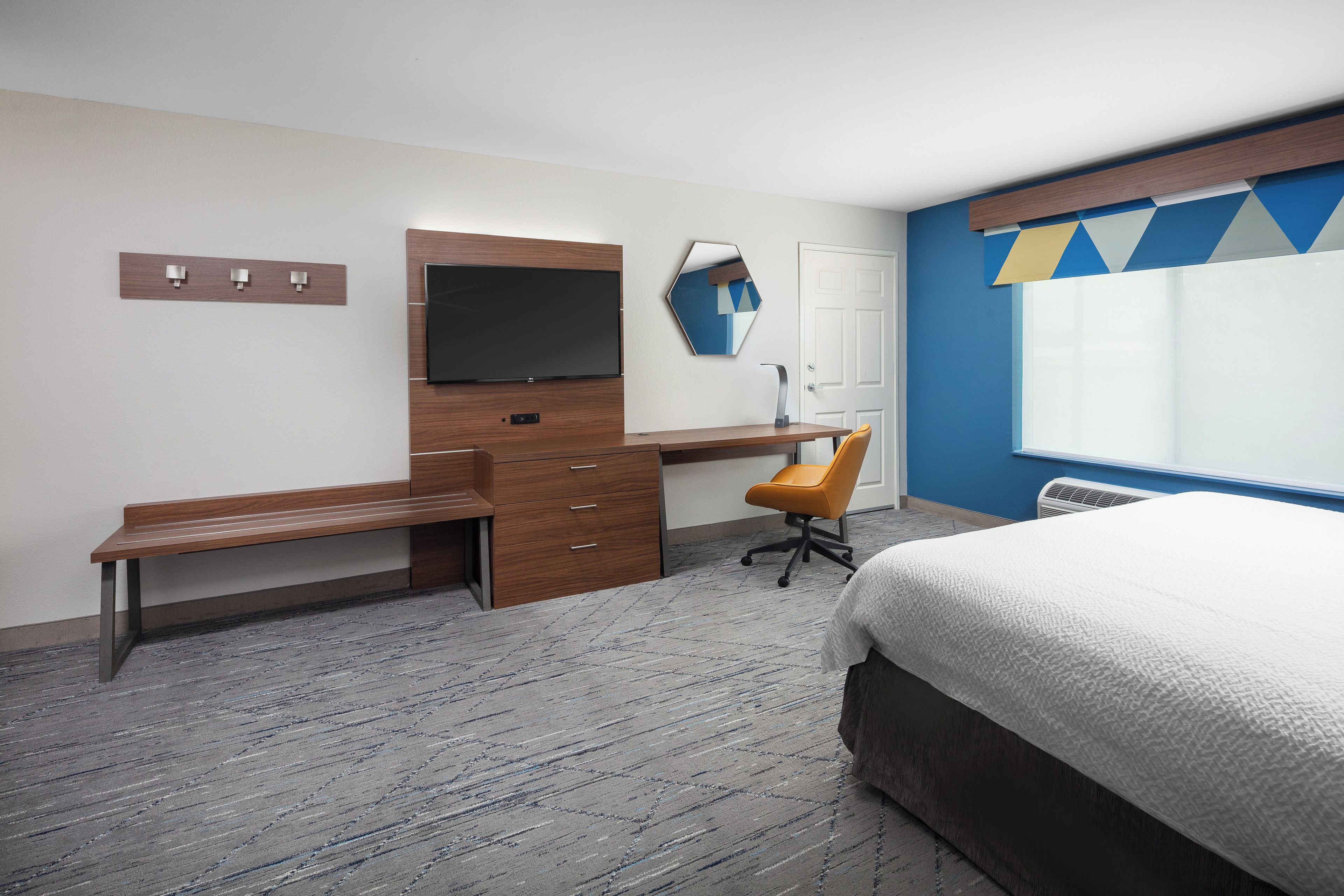 Habitación de hotel con cama, escritorio con silla, televisor de pantalla plana, espejo y ventana con cortina geométrica.